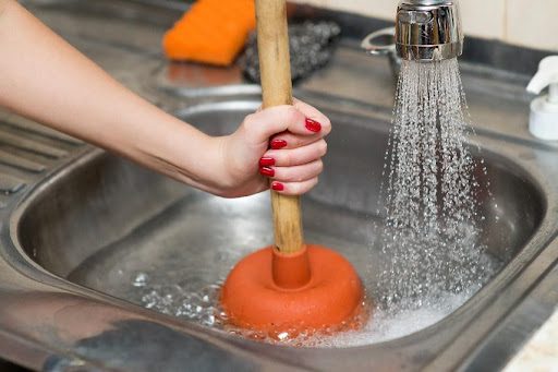 A female homeowner in Highland, IL, uses a plunger on the drain in her kitchen sink as water is running.